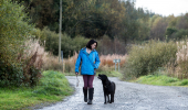 A woman in a blue jacket walks her black lab down a paved forest road