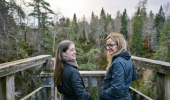 Two young women in blue jackets stand, smiling, on the wooden viewing platform overlooking forest of Douglas fir trees and Plodda Falls, Glen Affric