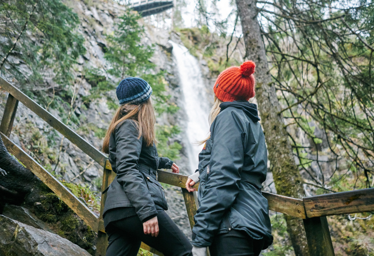 Two young women, one with orange woollen hat, other with blue striped woollen hat, look up from wooden viewing platform to high point of Plodda Falls waterfall as it roars down rockface, Glen Affric