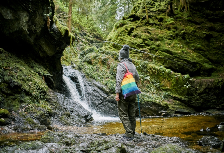  In a shady woodland glen, a female hiker, wearing grey jacket and hat, and carrying rainbow coloured bag, gazes at waterfall spilling over moss covered rocks into stream below, Puck's Glen, near Dunoon