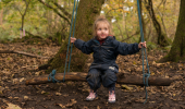 Young girl in blue wet weather gear sits on swing made of log, in Rannoch Wood, Renfrewshire Woods near Johnstone