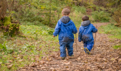 Two young boys in blue wet weather gear, each holding a stick, walk along forest path strewn with leaves, Rannoch Wood, Renfrewshire Woods, near Johnstone