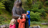 Three children in colourful jackets standing by a large wooden sculpture of a bear with trees behind.