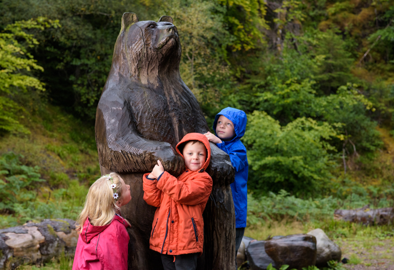 Three children in colourful jackets standing by a large wooden sculpture of a bear with trees behind.