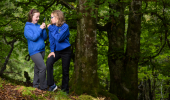 Two girls in blue jumpers laughing while taking a photo stood in a forest.