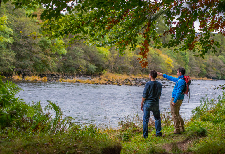 Two men standing on grassy river bank pointing across to trees on the far bank.