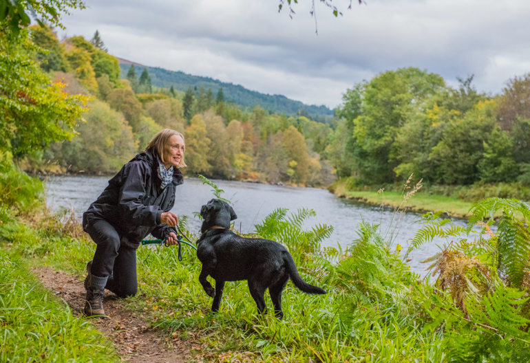 Woman kneeling down to play with black dog on small path beside a river with trees on each bank.