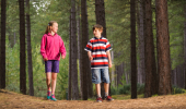 Girl and boy standing in a forest of tall trees on ground covered in brown pine needles.