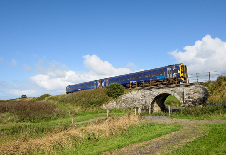 A blue train passes above a stone archway bridge alongside grass-lined pathways