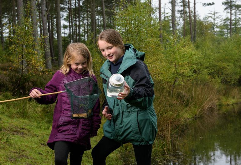 Two young girls with net and jar at the dipping pond, Skelbo Wood with forest and loch in background, near Dornoch, Sutherland