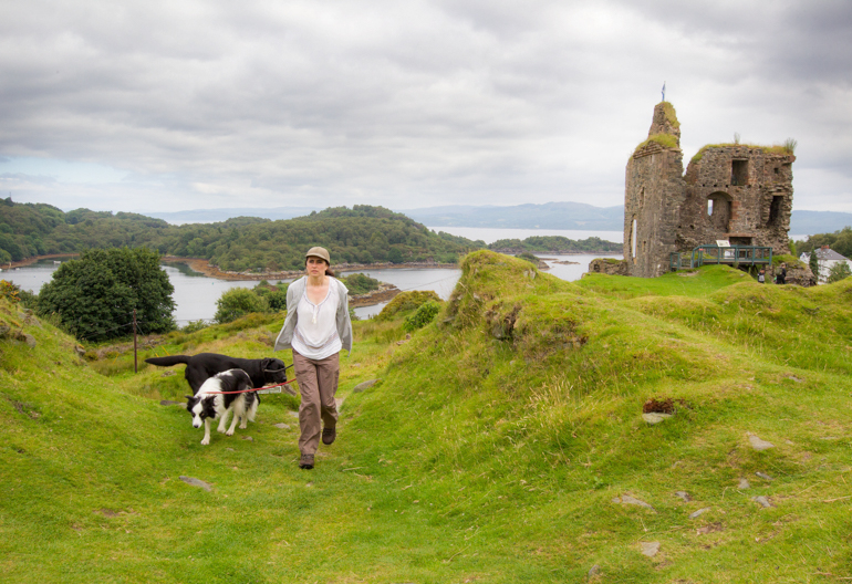 A woman walks two dogs along a grassy path next to the ruins of a small castle
