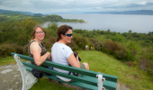 Two women sitting on a bench enjoying a view across woodland and a sea loch