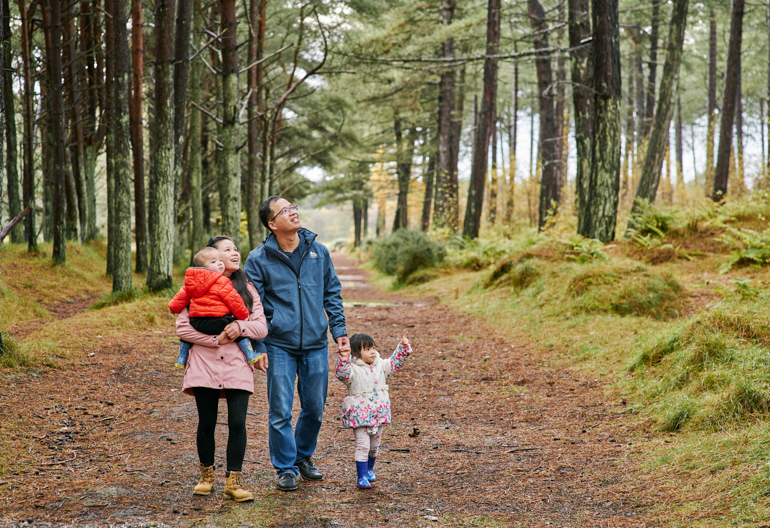 A family with a toddler and a baby look towards the treetops while walking along a woodland path