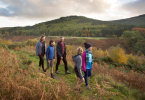A family of 5 walk along a grassy path with views onto autumnal conifer forest on the far side of a valley