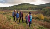 A family of 5 walk along a grassy path with views onto autumnal conifer forest on the far side of a valley