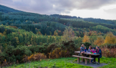 A family of 5 sit at a picnic bench looking across a valley towards thick conifer forest