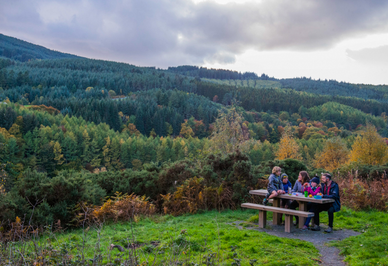 A family of 5 sit at a picnic bench looking across a valley towards thick conifer forest