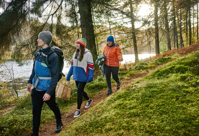 A man in a blue jaket, a woman in a rainbow hat, and a woman in a red jacket walk down a narrow trail next to a calm lake with moss and grass on the ground