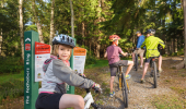 Girl on mountain bike by a signpost in a forest with family on bikes ahead.