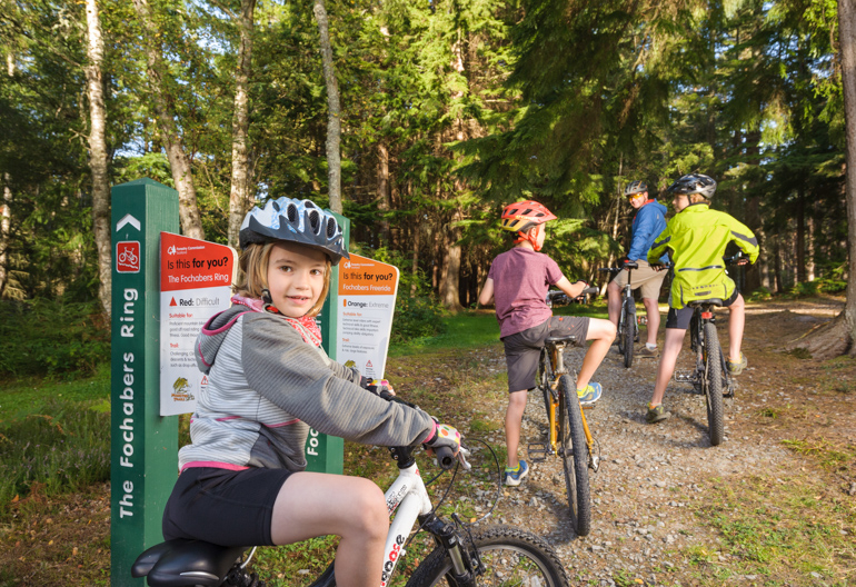 Girl on mountain bike by a signpost in a forest with family on bikes ahead.
