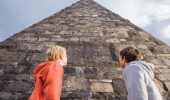 Two boys loking straight up at a dark stone pyramid and blue sky.