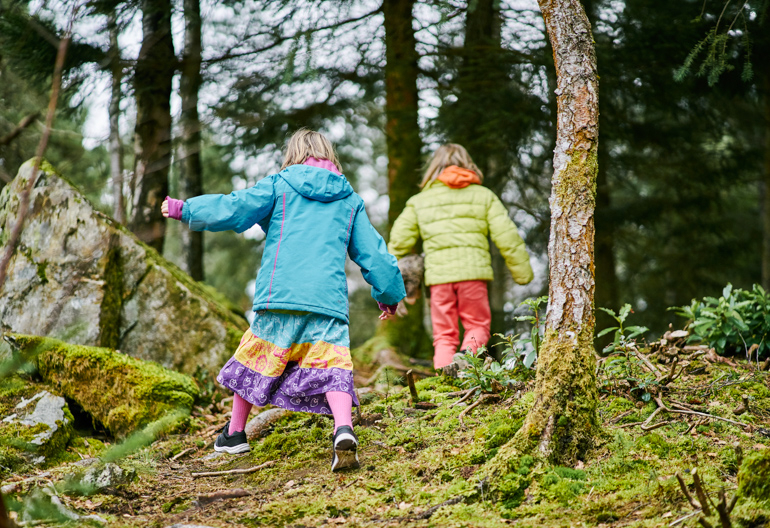 Rear view of two young girls, one in blue jacket, one in green, climbing up moss covered slope in forest, Faskally, near Pitlochry