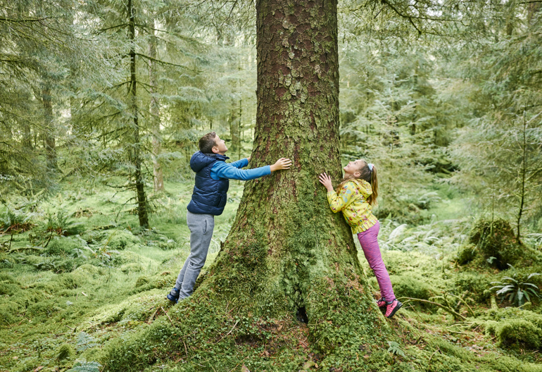 Boy and girl lean against a large tree covered in green moss from either side.