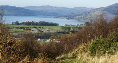 View down grassy track surrounded by scrub to green fields, open water and distant hills beyond on a bright, clear day.