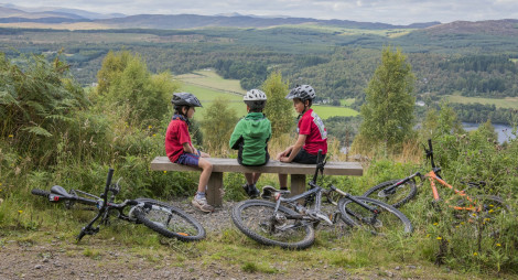 Three young boy cyclists sit on wooden bench high up in Balnain, with views overlooking Loch Ness and hills beyond