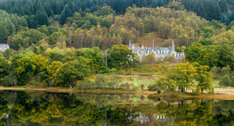 Large turreted house situated within green trees on the banks of a still loch.