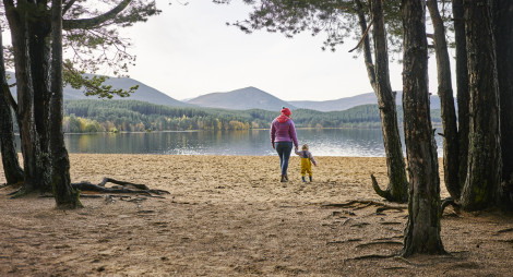 Woman and child walk on beach with trees around and on far shore of loch, and distant hills beyond.