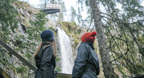 Two young women, one with orange woollen hat, other with blue striped woollen hat, look up from wooden viewing platform to high point of Plodda Falls waterfall as it roars down rockface, Glen Affric