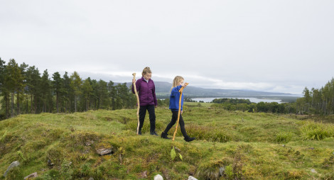 Two young girls walking beside broch remains on knoll in Skelbo Wood with forest and loch in background, near Dornoch, Sutherland