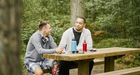 Two men (one with prosthetic leg) sit together chatting at picnic table, on woodland trail, Devilla Forest, near Kincardine