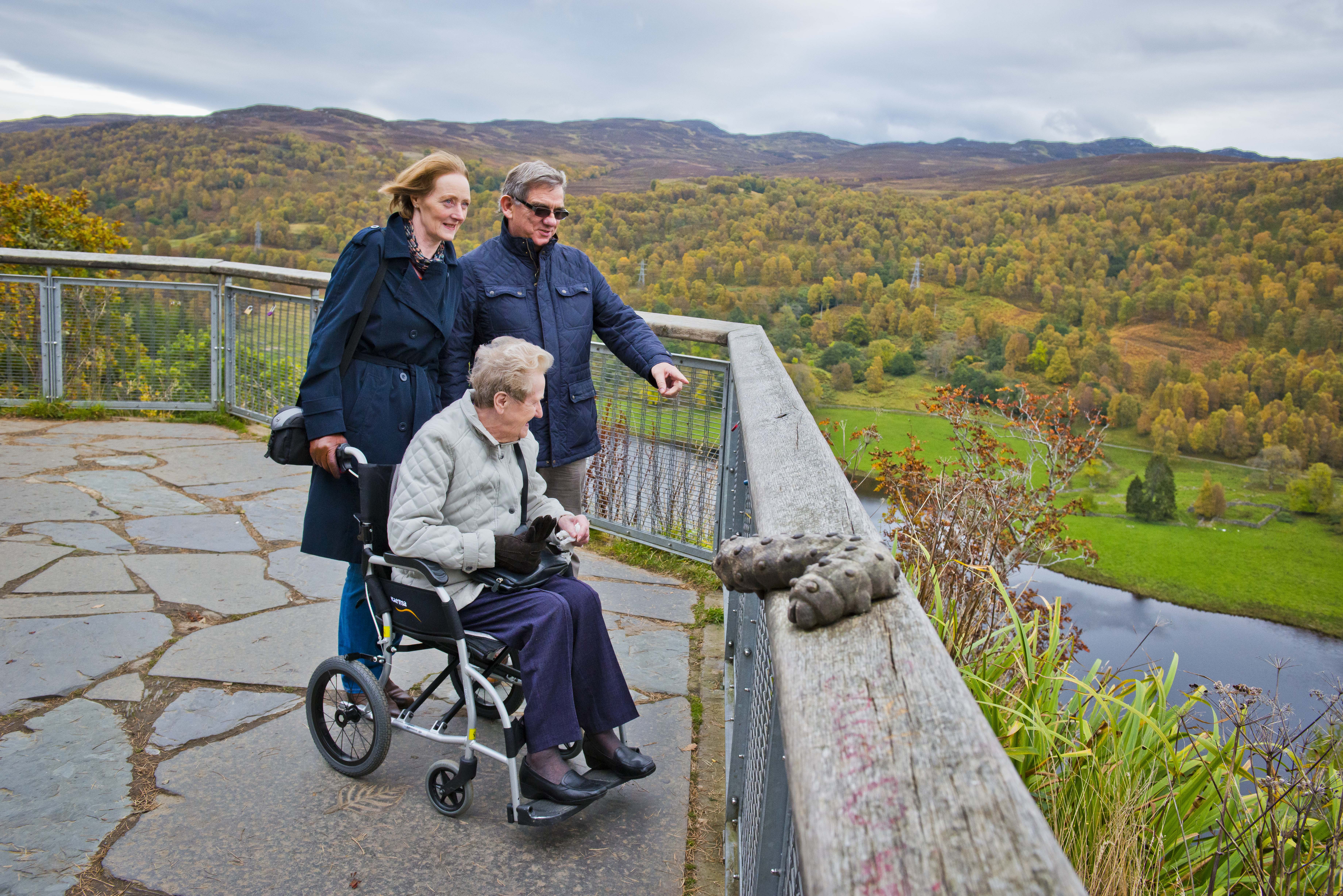 A women in a wheelchair pushed by another woman and a man look from the viewpoint at Queen's View Visitor Centre