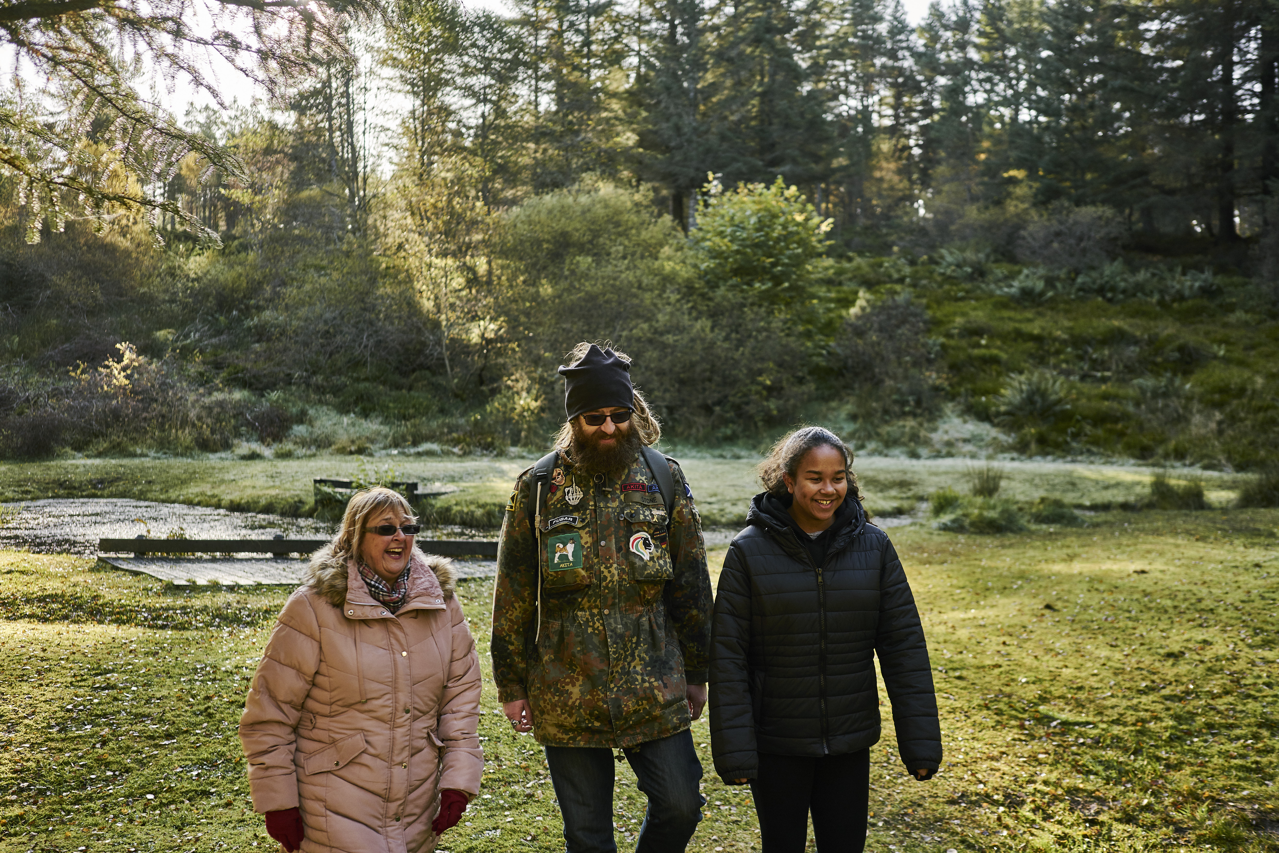 Teenage girl, man in combat jacket and dreadlocks and woman walking together with pond behind them, woodland trail, Aldie Burn forest, near Tain.