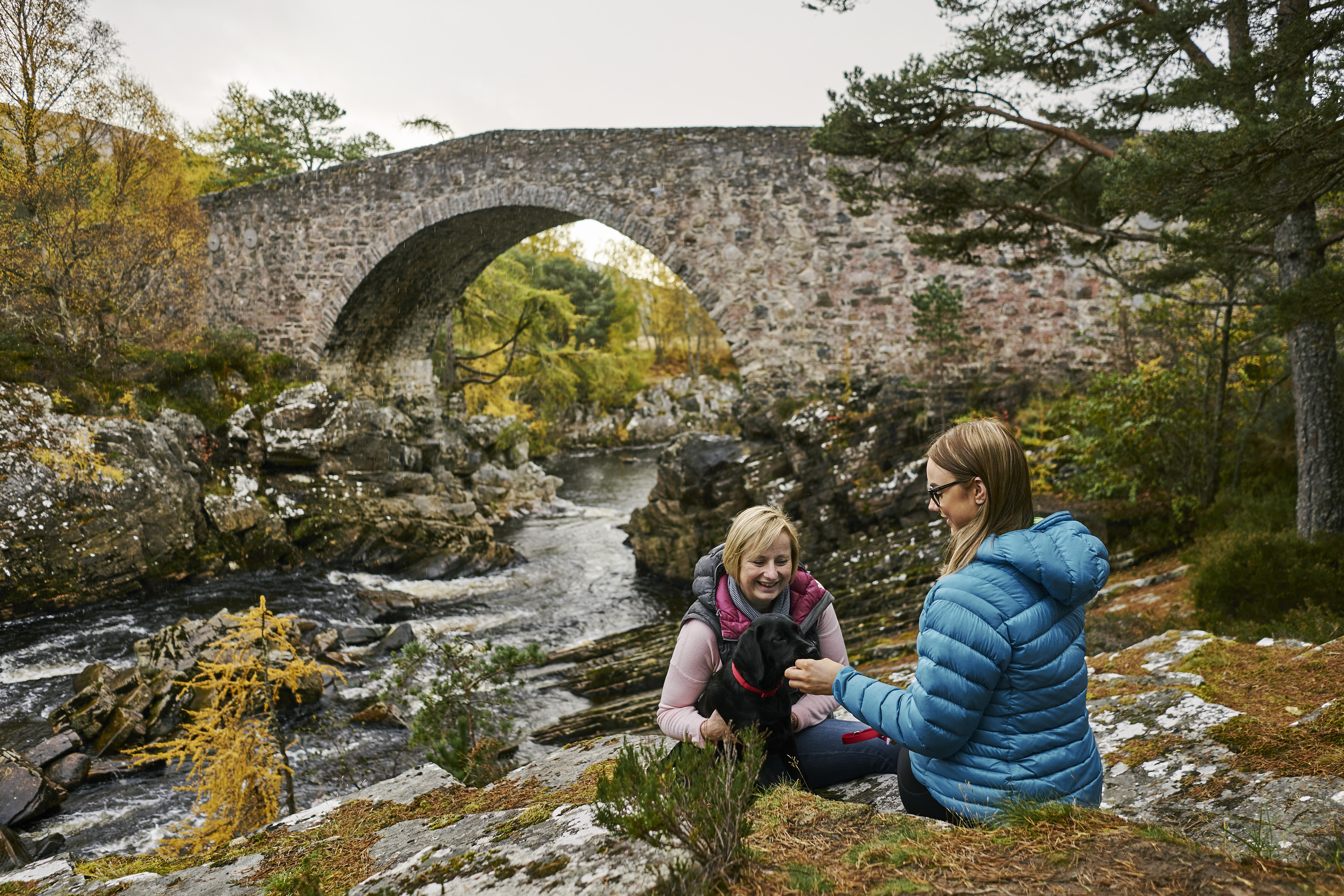 Two women sitting along the river bank with their dog, a stone bridge in the background, Little Garve