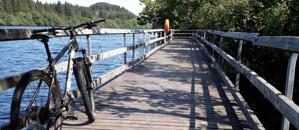 A wooden walkway over a body of water with trees beyond and two bikes in the foreground