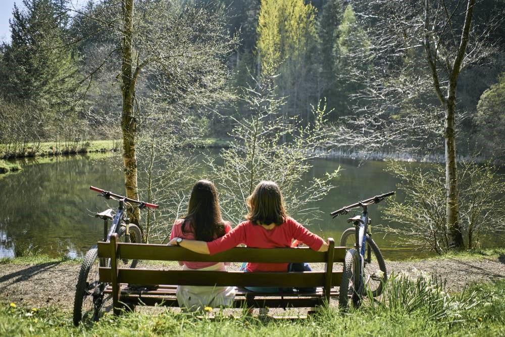 View to the rear of a bench with two people sat facing away from camera, bikes on either side of them, towards a body of water with tall trees beyond