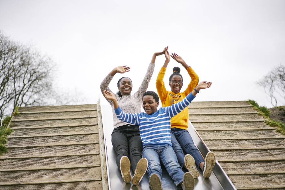 Three people smiling and laughing on a very wide playground slide or chute