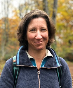 Woman in fleece wearing a rucksack smiling at camera with trees behind