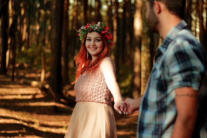 Red haired woman in dress smiling and holding out hand to man in checked shirt, all in forest surrounding