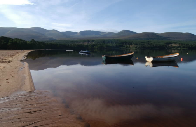 Two rowing boats moored near the shore of a still loch with tress on the distant shore and blue sky above