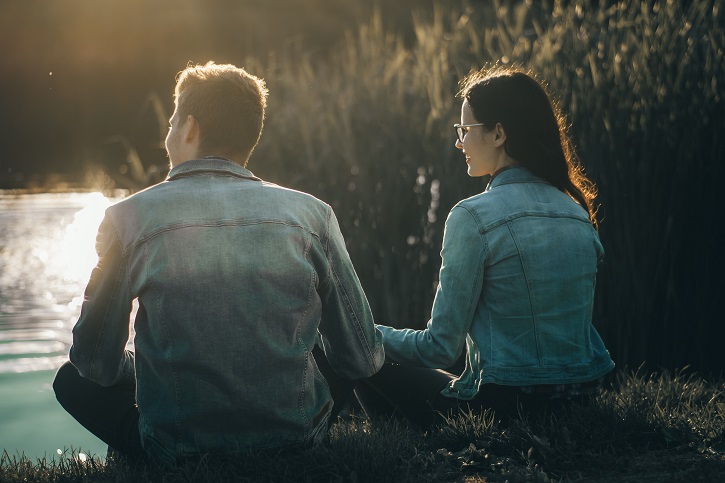 Man and woman both wearing denim jackets sat on grassy bank overlooking the landscape