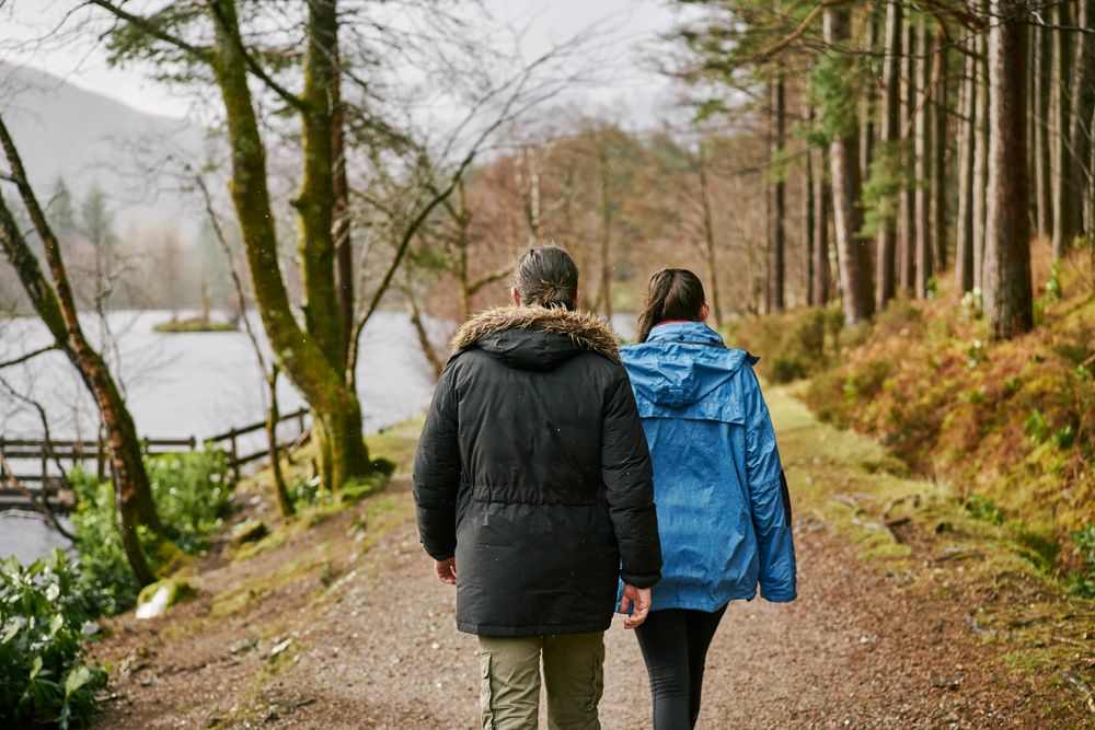 Rear view of young man in black jacket and young woman in blue jacket walking on path along banks of Glencoe Lochan after the rain, with tall conifers flanking the path and misty mountain range in the background