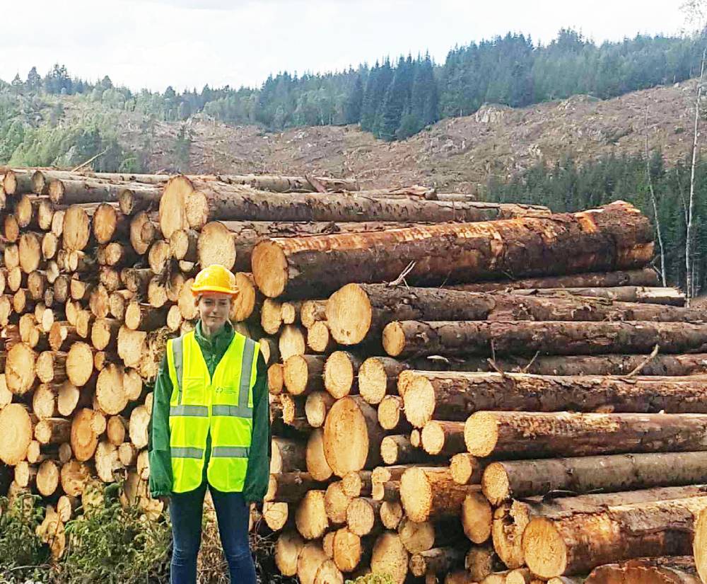 Woman in hi-vis vest and hard hat standing in front of pile of felled timber logs with forested hillside in background