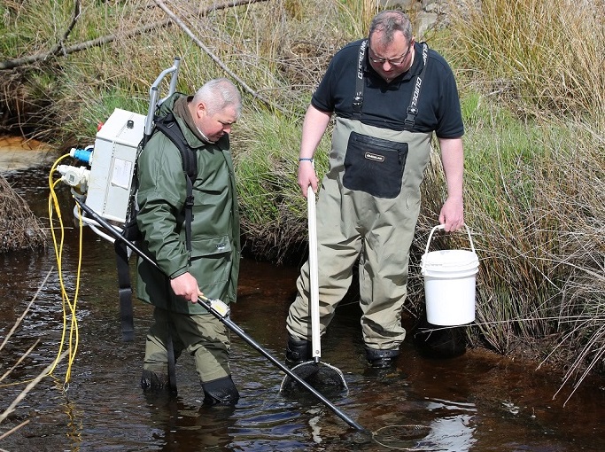 Two men in waders standing in a stream with fishing equipment