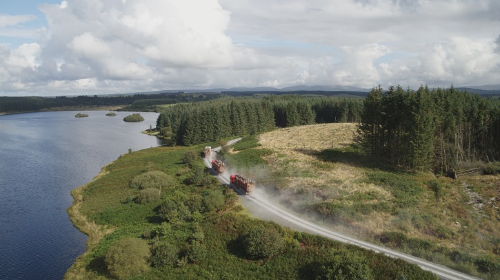 Aerial view of three large lorries with timber driving on a dirt road alongside a loch