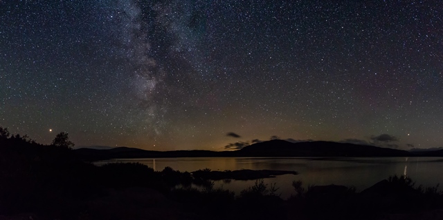 Photo at night of landscape showing stars and the silhouette of a distant forest with loch near at hand