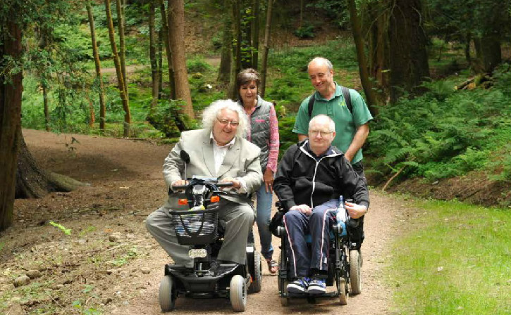 Four adults on a woodland road surrounded by trees, two on mobility scooters and two walking
